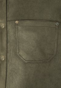 Grüner Wildlederstoff mit einer quadratischen Tasche, die sichtbare Nähte und Metallelemente aufweist. Die Textur wirkt weich und hat eine matte Oberfläche.