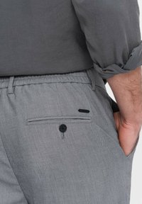 Man klädd i grå texturerade byxor med bakficka med knapp och en mörkgrå skjorta med uppkavlade ärmar, hand i framfickan.
