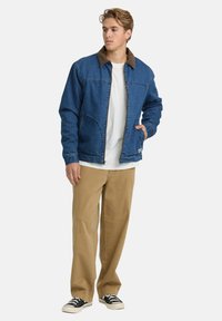 Dżinsowa kurtka z brązowym kołnierzem z sztruksu, z przodu na zamek, z dwiema kieszeniami bocznymi, noszona na białej koszulce i zestawiona z beżowymi spodniami i czarnymi butami.