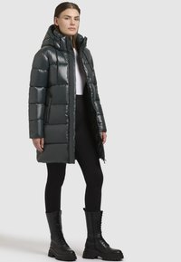 Gevoerde, lange jas in donkergroen, met een glanzende afwerking, afneembare capuchon en ritsen aan de voorkant. Draag het met zwarte leggings en knielange veterschoenen.