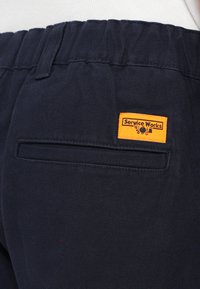 Granatowe spodnie z elastycznym pasem, posiadające tylną kieszeń oraz żółtą metkę "Service Works" przyszytą po boku.
