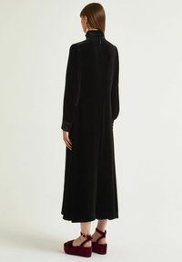 Abito lungo in velluto nero con collo alto e maniche lunghe, caratterizzato da una texture morbida. Abbinato a sandali platform color borgogna.