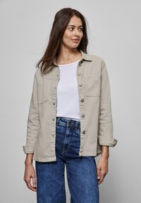 Beige knäppjacka med två framfickor, långa ärmar och lätt textur; bärs över en vit t-shirt och matchas med blå jeans.