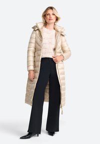 Langer, cremfarbener Daunenmantel mit horizontaler Steppung und Kapuze. Verfügt über einen Frontreißverschluss und eine Schnur. Kombiniert mit einem leichten Strickpullover und schwarzen Hosen.
