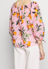 Donna che indossa un blouse rosa scoperto sulle spalle con stampa di frutti arancioni e foglie verdi, abbinato a pantaloni bianchi, vista da dietro.