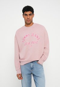 Rozā kapuce ar apaļu kakla izgriezumu, ar baltu izšūtu tekstu "TOMMY JEANS NEW YORK CITY." Nēsāta ar gaiši zilām džinsu biksēm.