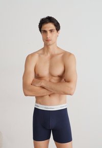 Boxeri boxer albaștri închis cu un brâu simplu alb, realizați dintr-un material moale, cu un design ajustat și o textură netedă. Modelul stă cu brațele încrucișate.