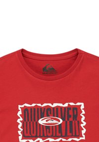 Camiseta de algodón roja con cuello redondo. Presenta un diseño gráfico de logo en blanco y azul en la parte delantera, contorneado con un patrón en zigzag.