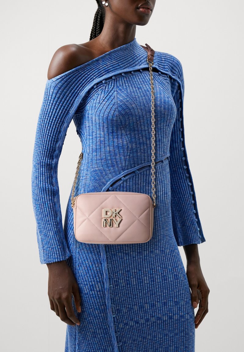 Femme portant une robe en maille côtelée bleue avec une épaule découverte, portant un petit sac bandoulière matelassé DKNY rose clair avec une chaîne dorée.