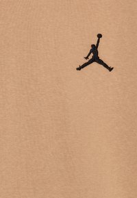 Tela de algodón marrón claro con un logotipo de Jumpman bordado en negro en una pose dinámica, que destaca por su textura suave y diseño minimalista.