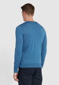 Niebieski sweter z długimi rękawami i okrągłym dekoltem, z teksturowanym splotem oraz ściągaczami przy mankietach i u dołu. Noszony z ciemnymi dżinsami.