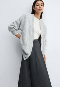 Jeune femme portant un cardigan ample gris clair par-dessus un haut blanc et une jupe plissée taille haute gris foncé, posant devant un fond uni.