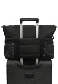 Čierna prešívaná taška na príručnú batožinu upevnená na čiernom kufri na kolieskach. Obsahuje hornú rukoväť, zipsy a nastaviteľné popruhy s vzorovanými akcentmi.