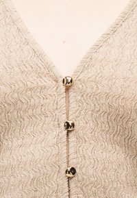 Μπεζ υφασμάτινο πλεκτό ένδυμα με βαθύ V-λαιμόκοψη, κλειστό με τρία γυαλιστερά στρογγυλά χρυσά κουμπιά στο μπροστινό μέρος.