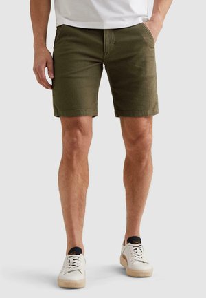 Mann trägt olivgrüne, strukturierte Shorts, weißes T-Shirt und weiße Turnschuhe, steht mit den Händen in den Taschen.