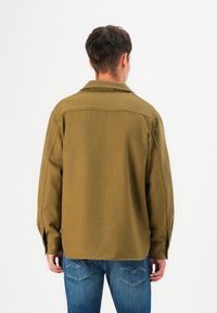 Olivengrøn skjortejakke med lange ærmer, krave og rygpilsdetail. Båret over blå jeans med karakteristisk lommebroderi.