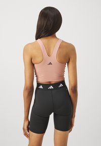 Roza sportska majica s racerback dizajnom i crnim Adidas logotipom, u paru s crnim hlačama visokog struka koje imaju uzorak na pojasu.