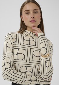 Dlhý rukáv, rolákový top v krémovej farbe s čiernymi geometrickými vzormi. Vyznačuje sa textúrovanou tkaninou a zlatým detailom na lakti.