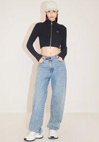 Top corto de punto negro con cremallera, pantalones vaqueros de pierna ancha en azul claro y zapatillas blancas. La gorra es de un beige claro con un diseño simple.