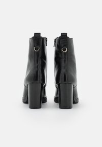 Botas de cuero negras hasta el tobillo con tacones en bloque, anillos de metal plateado en los lazos traseros, de pie sobre un fondo blanco mirando hacia atrás.