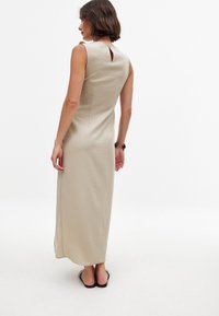 Beżowa, bezrękawnikowa maxi sukienka z dekoltem w kształcie łezki, bocznymi rozporkami i fakturowanym materiałem. Noszona z czarnymi klapkami, minimalne detale.