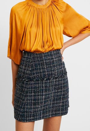 Vrouw draagt een geplooide, loszittende oranje blouse die is ingestopt in een high-waisted donker geruite rok met franjes.