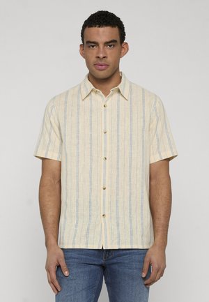 Mand iført en kortærmet, beige skjorte med knapper og lodrette blå striber samt blå jeans, stående mod en ensfarvet lys baggrund.