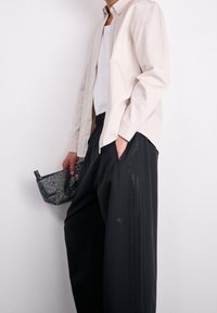 Světlý béžový košilový top, bílý podvlékací triko, černé kalhoty s díly z síťoviny po stranách, držící černou síťovanou tašku s rukojetí.