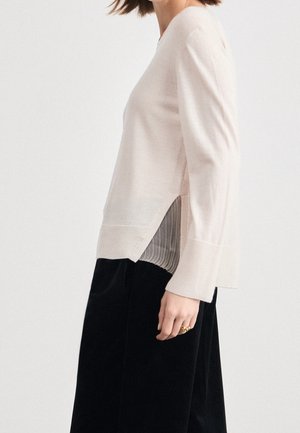 Frau trägt einen hellbeigen Pullover mit langen Ärmeln und seitlichem Schlitz über einem gestreiften Hemd, kombiniert mit schwarzen Hosen und goldenen Ringen an der Hand.