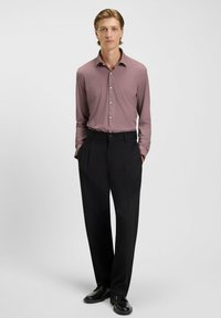 Junger Mann steht mit den Händen in den Taschen, trägt ein rot gemustertes Langarmhemd, schwarze Hosen und schwarze Lederschuhe.