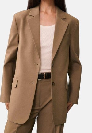 Vrouw draagt een op maat gemaakte camelbruine blazer en bijpassende broek met een zwarte riem en witte geribbelde tanktop.