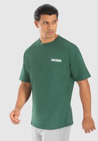Camiseta de algodón verde con mangas cortas, escote redondo y logo blanco "SMILODOX" en el lado izquierdo del pecho. Corte relajado, textura suave.