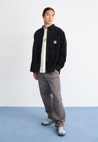 Jeune homme debout, une main dans la poche de son pantalon cargo gris, portant une chemise noire boutonnée sur un t-shirt blanc à motifs et des baskets grises.