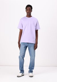 Camiseta corta de color púrpura claro, de corte relajado, combinada con jeans azules. Zapatillas deportivas blancas con rayas negras. Diseño simple, sin patrones.
