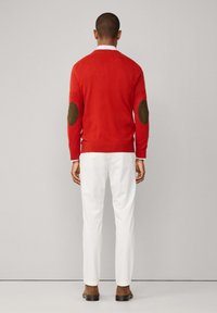 Muž v červeném svetru s hnědými loketními výztuhami, bílých kalhotách a hnědých botách, stojící čelem k jednoduché světle šedé zdi.