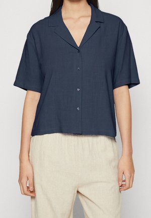 Vrouw die een marineblauw overhemdd met korte mouwen, kraag en lichte beige losse broek draagt.