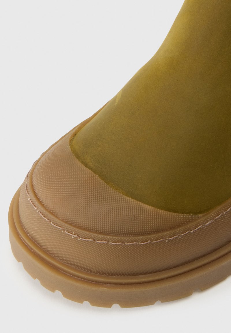 Botte en caoutchouc vert olive avec une semelle brune texturée. Bout arrondi, accents cousus et partie supérieure lisse.
