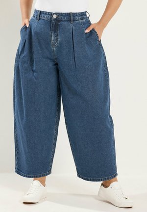 Pantalones vaqueros azules de pierna ancha con cintura alta y pliegues delanteros, combinados con zapatillas blancas y una camiseta blanca, manos en los bolsillos.