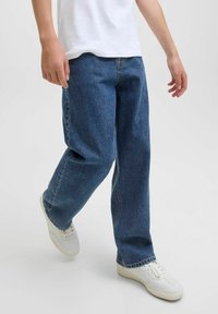 Jean en denim bleu à coupe décontractée avec une texture légèrement décolorée, associés à des baskets blanches. Les jeans présentent un design classique à cinq poches.