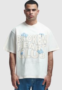 Fehér pamut póló, amelyen a "BROKEN HEART CLUB" felirat olvasható vastag fekete és kék betűkkel, szív grafikákkal. Laza fazon, rövid ujjú.