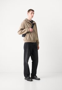 Beige lett jakke med elastisk kant, hette og grønn logo; sammen med svarte jeans og svarte sko. Mannen holder en liten svart veske.