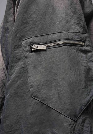 Tissu gris foncé avec une surface texturée. Comprend une poche zippée avec des éléments en métal et un patch triangulaire à proximité.