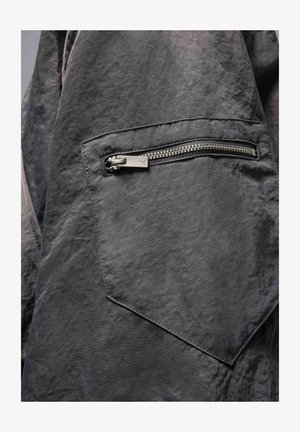 Dark gray fabric with a textured surface. Features a zippered pocket with metal hardware and a triangular patch adjacent to it.