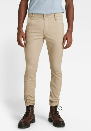 Mężczyzna w beżowych obcisłych spodniach i ciemnobrązowych sznurowanych butach, stojący z rękami swobodnie opuszczonymi wzdłuż ciała.