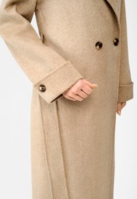 Beige wollen jas met een dubbelbreasted ontwerp, brede revers en opvallende bruine knopen. Heeft een zachte textuur en een taillering.