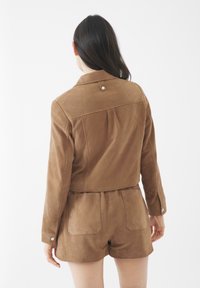 Giacca in suede marrone con un design corto, caratterizzata da un colletto con bottoni, pieghe posteriori e cuciture visibili, abbinata a pantaloni corti coordinati.