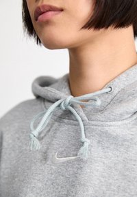 Grå bomullshoodie med en framficka, med ljusblå snörning och en liten broderad logotyp på bröstet. Texturerad tyg synlig.