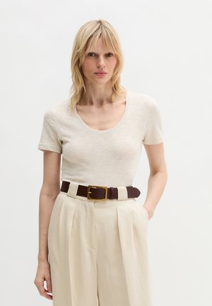 Blonde vrouw met een crèmekleurig T-shirt met ronde hals, ingestopt in een hoge taille plissé beige broek met een donkerbruine riem.