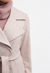 Mujer vestida con un abrigo de lana beige con cinturón sobre un suéter de cuello alto rosa empolvado, contra un fondo blanco.