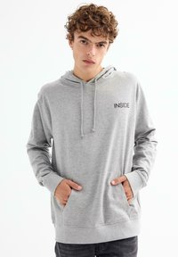 Sudadera con capucha gris, fabricada en tejido suave, que incluye un bolsillo frontal y el logo "INSIDE" en el pecho. Mangas largas y capucha ajustable con cordón.
